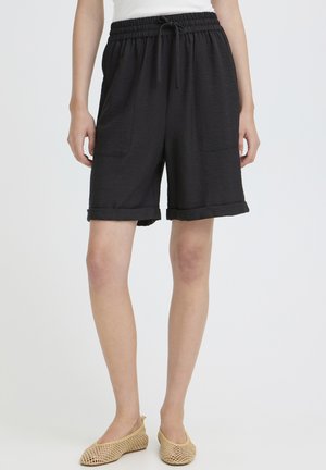 Nő fekete, elasztikus derékpántú, első zsebes rövidnadrágban és bézs színű hálós slip-on cipőben, egyszerű háttér előtt áll.