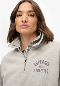 Mujer con cabello castaño hasta los hombros que lleva una sudadera gris con cremallera hasta el cuarto, con "Superdry Athletics" y un pequeño emblema de la bandera estadounidense.