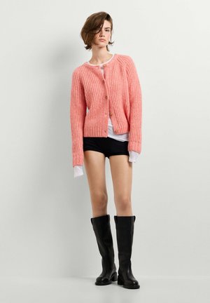 Junge Person trägt eine rosafarbene grob gestrickte Strickjacke, weißes Hemd, schwarze Shorts und hohe schwarze Lederstiefel und steht vor einem schlichten hellen Hintergrund.