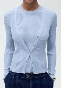 Cárdigan de canalé azul claro con mangas largas, escote en V profundo y seis botones en la parte delantera. Combinado con pantalones de mezclilla oscuros de tiro alto.