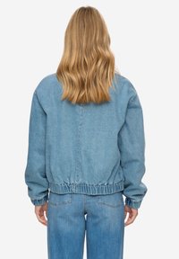 Modrá džínová bunda s elastickým lemem a manžetami, s volným střihem a jednoduchým, čistým designem. Nošena přes odpovídající džínové kalhoty.