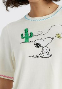 Gebreid sweater in crème met lichtblauwe en roze accenten, voorzien van een cactus en Snoopy ontwerp in zwarte en groene borduursels.