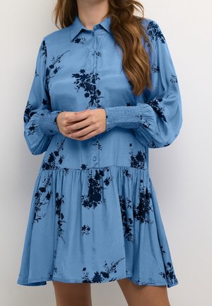 Vrouw die een loszittende blauwe jurk draagt met zwarte bloemmotieven, lange opgeblazen mouwen met gerimpelde manchetten en een knoopsluiting aan de voorkant.