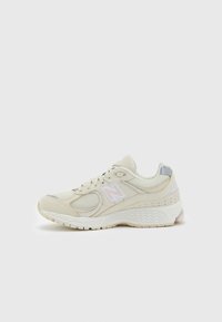 Beige athletic shoe with mesh, leather, and synthetic accents. Features a rounded toe, lace-up design, and pink and gray detailing on the heel.