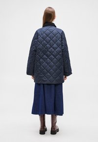 Tengerészkék steppelt dzseki álló gallérral, bő fazonnal és leejtett vállakkal, hosszú farmer szoknyával és térdig érő barna csizmával párosítva.