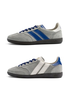 Deux baskets en daim gris avec des semelles noires ; la chaussure du dessus a des bandes et des lacets bleus, la chaussure du dessous a des bandes blanches et des accents bleus au talon.