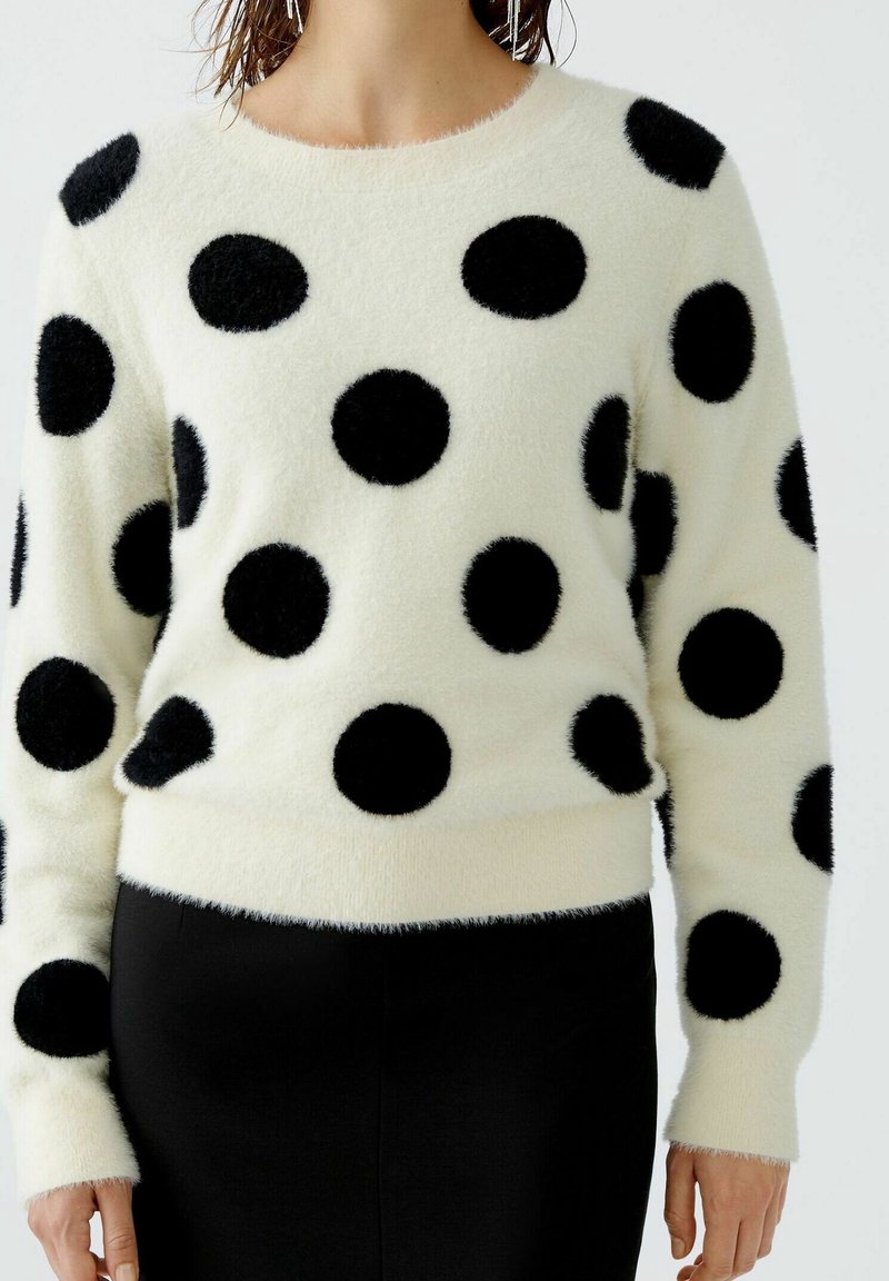 Pull blanc duveteux avec de grands pois noirs porté avec un pantalon noir, montré du cou aux hanches.