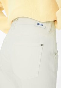 Primo piano della tasca posteriore di jeans bianchi con patch del logo Brax indossati con maglione giallo chiaro.