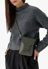 Borsa a tracolla verde in pelle con finitura testurizzata, chiusura a linguetta, hardware in metallo e logo del brand. Indossata su un maglione grigio.