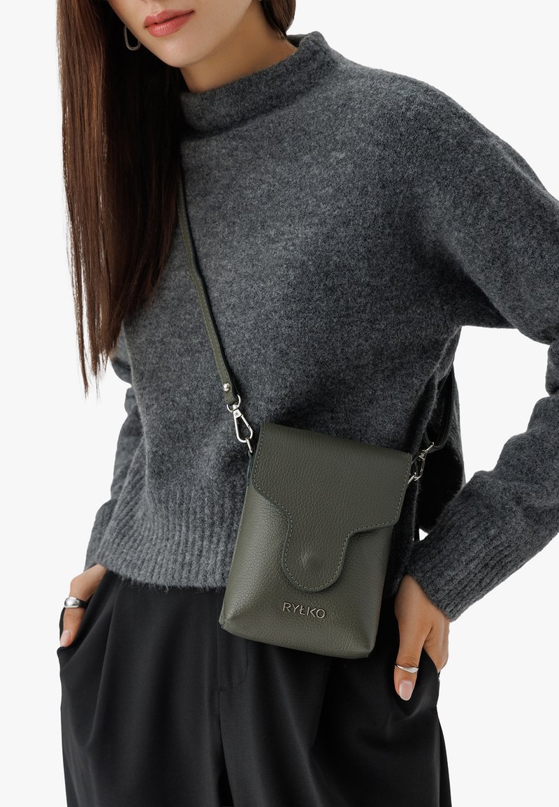 Borsa a tracolla verde in pelle con finitura testurizzata, chiusura a linguetta, hardware in metallo e logo del brand. Indossata su un maglione grigio.