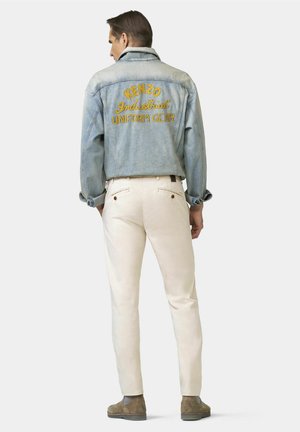 Mann von hinten, trägt eine hellblaue Jeansjacke mit gelbem Schriftzug "KENZO Industrial Uniform Gear", beige Hose und braune Wildlederschuhe.