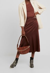 Gonna midi in satin bordeaux con design drappeggiato, abbinata a un blazer beige e una borsa a tracolla con stampa serpente marrone, indossata con stivaletti neri con tacco.