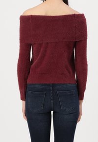 Pull bordeaux à épaules dénudées en tissu texturé. Avec des manches longues et une taille ajustée, associé à un jean skinny bleu foncé.