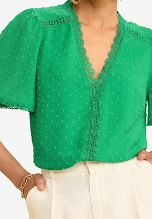 Groene blouse met textuur, V-hals afgezet met kant en korte pofmouwen, gecombineerd met crèmekleurige hoge taillebroek en gouden armbanden.