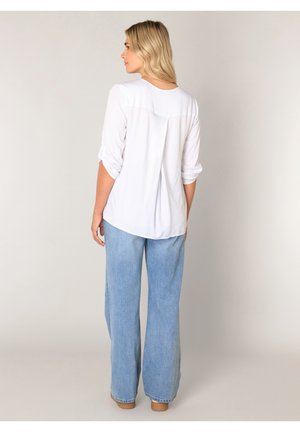 Woman with blonde hair wearing a white long-sleeve blouse and light blue wide-leg jeans, standing facing away against plain background.