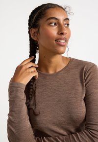 Top a maniche lunghe di colore marrone, realizzato in un tessuto texturizzato, con scollatura rotonda e maniche aderenti. La persona ha i capelli intrecciati e sta toccando i suoi capelli.