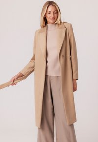 Beżowy wełniany płaszcz o dopasowanym kroju, z klapami i pojedynczym zapięciem na guzik. Prezentowany na tle jasnobeżowego swetra dzianinowego i szerokich spodni.