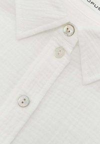 Camicia bianca con texture e colletto a punta, dotata di tre bottoni rotondi simili a perle nella parte frontale e un sottile motivo a griglia sul tessuto.