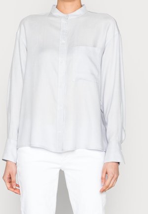 Kobieta nosząca luźną, długą koszulę na guziki w jasnoszarym kolorze z kieszenią na piersi oraz białe spodnie.