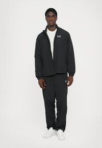 Black zip-up jacket with a textured fabric, featuring a standing collar and EA7 logo. Paired with matching black pants and white sneakers.