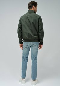 Chaqueta bomber de color verde oscuro con puños y cintura elásticos, combinada con jeans de color azul claro y zapatillas blancas, vista desde la parte de atrás.
