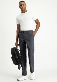 Pantalones grises con cierre de botón, de textura suave, combinados con una camiseta blanca ajustada y una mochila negra. Las zapatillas blancas completan el look.