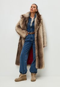 Bruin nepbontjack met een lange snit, gelaagd over een blauwe denim jumpsuit. Beigekleurige enkellaarsjes maken de outfit compleet. Glad textuur overal.
