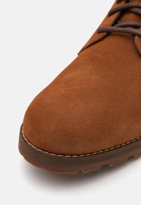 Chaussure en cuir suédé marron avec un bout arrondi, dotée de lacets marron et d'une semelle en caoutchouc durable, présentant une texture lisse et douce.