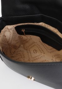 Interiér černé kožené kabelky s béžovým podšívkou, obsahující zipovou kapsu a potištěný design loga. Hladká textura a zaoblený tvar.