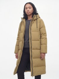 Femme portant un long manteau matelassé beige avec capuche, non fermé, par-dessus un pull gris foncé et un pantalon noir, debout devant un fond blanc.