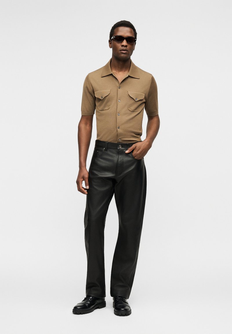 Homme portant des lunettes de soleil foncées, une chemise marron à manches courtes boutonnée, un pantalon en cuir noir et des chaussures noires, debout avec une main dans la poche.