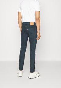 Mörkblå slim-fit jeans med en läderbaklapp och en röd etikett på fickan, kombinerat med en vit t-shirt och vita sneakers.
