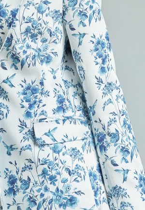 Jacke mit Blumenmuster in weißem Stoff mit blauen Blumen und Kolibris, mit einer einzelnen Taschen-Detail, glatte Textur.