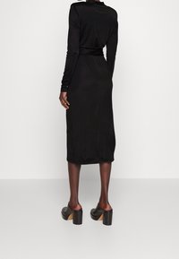 Schwarzes langärmliges Kleid aus weichem Stoff, mit einer taillierten Taille und einem Rock, der bis zur Mitte der Wade reicht. Kombiniert mit klobigen schwarzen High Heels.