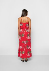 Vestido maxi vermelho com padrão floral, alças spaghetti ajustáveis, design com costas franzidas e silhueta fluida. Sandálias pretas de salto bloco.