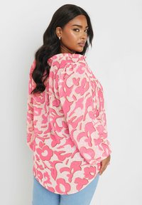 Camicia a maniche lunghe rosa con motivo floreale, caratterizzata da un collo e polsini arricciati e un orlo curvo. Tessuto morbido con una texture leggermente trasparente.