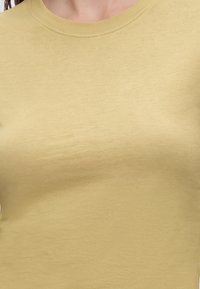 Κοντινό πλάνο ενός ατόμου που φοράει μια απλή ανοιχτόκίτρινη μπλούζα με στρογγυλή λαιμόκοψη, που δείχνει την υφή του υφάσματος και το ράψιμο στον γιακά.
