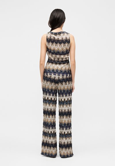 Γυναίκα στέκεται με την πλάτη, φοράει αμάνικο ολόσωμο με κυματιστές ρίγες σε μπεζ, μαύρες, λευκές και μπλε αποχρώσεις.