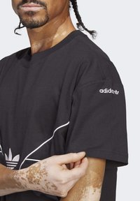 Man med flätat hår som bär en svart Adidas T-shirt, visar detalj på ärmen med vitt märke på överarmen och vita geometriska linjer på bröstet.