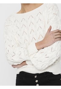 Witte gebreide trui met een open geweven patroon, brede bootnek en geribbelde manchetten en zoom. Model slaat armen voor zich.