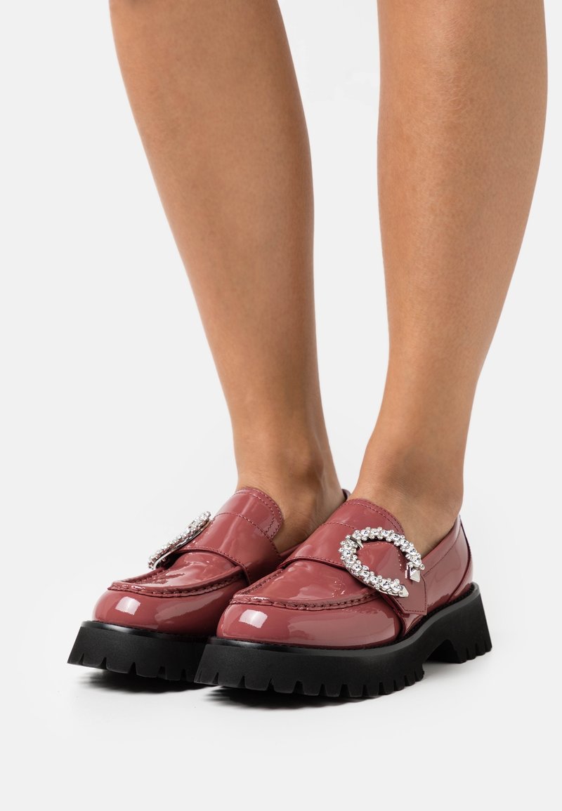 Mocassins en cuir verni de couleur rose profond avec une semelle noire épaisse, présentant un détail en métal orné de strass décoratifs à l'avant.