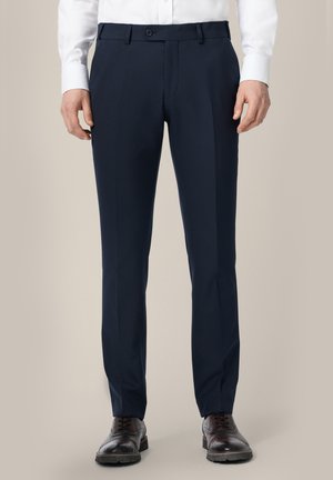 Mężczyzna w granatowych, dopasowanych eleganckich spodniach, białej koszuli i ciemnobrązowych skórzanych butach brogue, stojący na jednolitym tle.