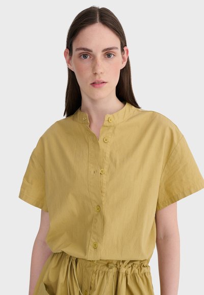 Jeune femme aux cheveux bruns raides portant une chemise moutarde à manches courtes avec boutons et un pantalon assorti à taille froncée.