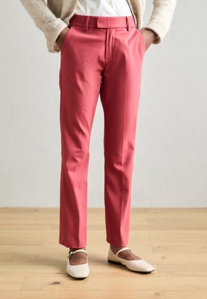 Woman wearing coral slim-fit trousers with side pockets, beige cardigan, white blouse, and white Mary Jane shoes standing on wooden floor.