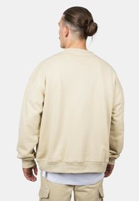 Felpa beige con vestibilità rilassata, polsini e orlo a costine, e una texture morbida, indossata sopra una t-shirt bianca e abbinata a pantaloni beige.