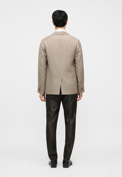 Μπεζ σακάκι προσαρμοσμένης ραφής με γιακά V, δύο κουμπιά και σχισμή πίσω, συνδυασμένο με καφέ παντελόνια με σχέδιο και μαύρα παπούτσια.