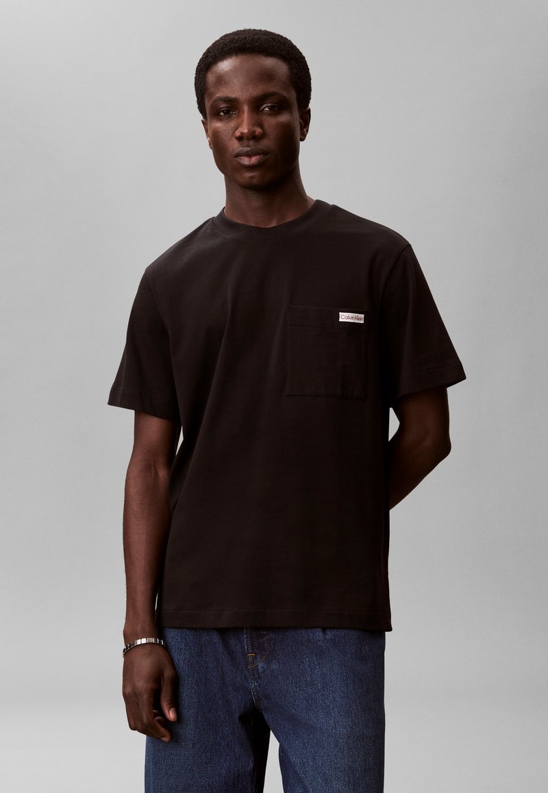 Νέος άντρας φοράει μαύρο κοντομάνικο T-shirt με τσέπη μπροστά και μπλε τζιν, στέκεται μπροστά σε απλό γκρι φόντο.