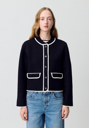 Veste noire coupée avec des finitions blanches, col rond et fermeture à boutons. Comprend deux poches avant. Portée sur un jean en denim bleu.