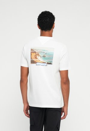 Fehér pamut póló, hátul grafikai nyomattal, amely egy tengerparti tájat ábrázol kék és bézs színekben, valamint egy "Tommy Hilfiger" logóval.