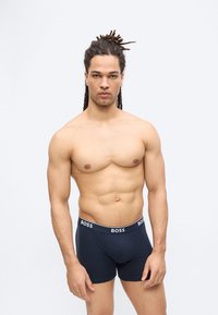 Μυώδης άνδρας με ντραντλόκς στέκεται αντικρίζοντας μπροστά φορώντας μαύρα ανδρικά εσώρουχα BOSS μπροστά σε λευκό απλό φόντο.